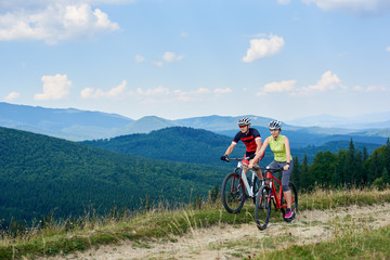 Young bikers tourists, man and woman in professional sportswear riding bikes down grassy field road under beautiful bright blue sky on magnificent mountain range background. Active lifestyle concept.