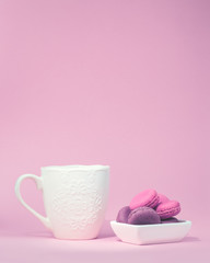 Several sweet tasty pink macarons in white bowl and white coffee mug on pink background with copy space closeup