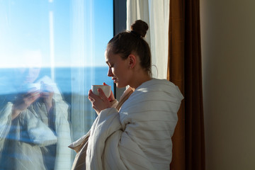 Pretty young woman wrapped in a blanket is standing near window and enjoying first morning coffee on sunshine. An early waking and the beginning of a new day