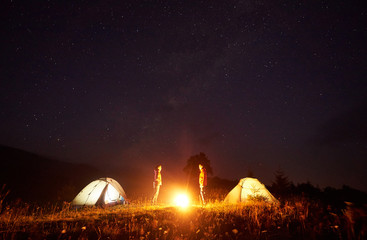 Night camping. Bright bonfire burning between two tourists, boy and girl standing opposite each other in front of illuminated tents under beautiful dark starry sky on distant hills background.