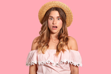 Portrait of shocked Caucasian woman wears straw hat and fashionable blouse, shows bare shoulders, isolated over pink background, surprised to have spoiled vacation. Omg and reaction concept.