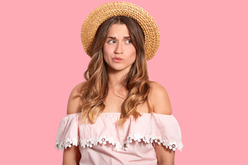 Thoughtful puzzled woman looks aside, has long wavy hair, wears summer straw hat and blouse, thinks about future vacation, isolated over pink background. People and facial expressions concept