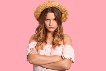Serious gloomy young cute woman with dissatisfied facial expression, keeps arms folded, looks from eyebrows, wears straw hat and blouse, isolated over pink background. Negative emotions concept