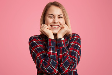 Close up shot of satisfied woman holds chin, smiles broadly, shows white perfect teeth, being in good mood, dressed casually, poses against pink background. Optimistic teenager expresses happiness