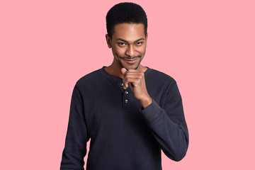 Obraz premium Indoor shot of handsome black young man holds chin, looks with pleased cunning facial expression, wears casual sweater, listens pleasant story from interlocutor, isolated over pink background