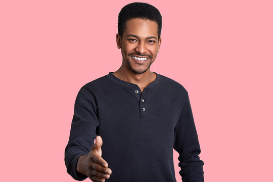 Positive Happy Black Man Gives Handshake, Greets With Friend, Dressed In Casual Jumper, Smiles Broadly, Has White Shining Teeth, Isolated Over Pink Background. Meeting Gesture. Body Language