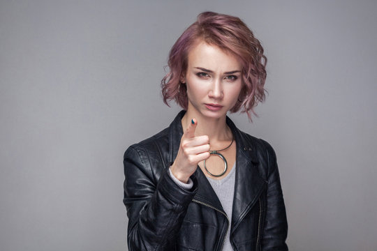 Portrait of caution serious beautiful girl with short hairstyle and makeup in casual style black leather jacket standing, warning and looking camera. indoor studio shot, isolated on grey background.
