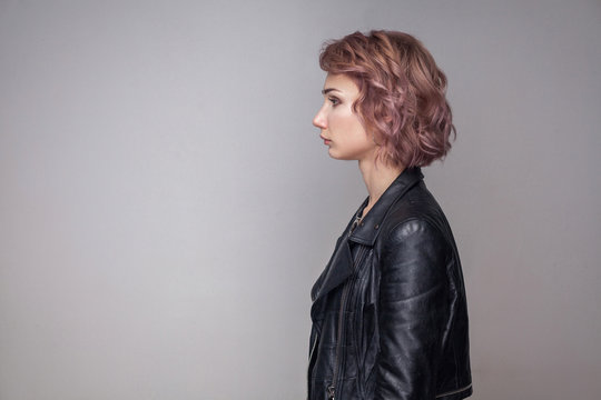 Profile side view portrait of serious beautiful girl with short hairstyle and makeup in casual style black leather jacket standing and looking aside. indoor studio shot, isolated on grey background.