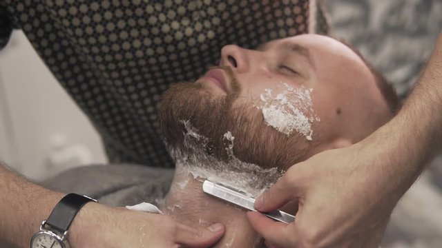 Barber shaving bearded man with straight razor in male salon. Male skin care and beard style concept in slow motion
