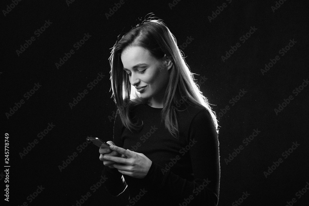 Wall mural black and white portrait of young woman looking at phone and smiling, isolated studio photo on dark 