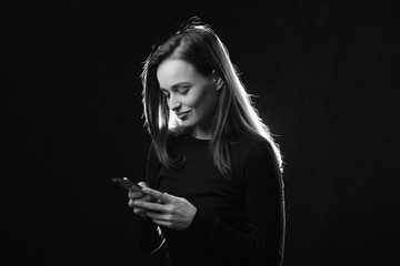 Black and white portrait of young woman looking at phone and smiling, isolated studio photo on dark background. Beautiful model with long hair using cell phone
