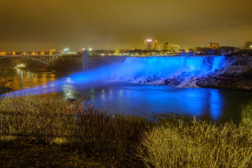 Naklejka na ściany i meble Scenic view of American falls and Bridal Veil Falls illuminated by blue nighttime illumination on Niagara river in Canada. Beautiful depressive winter look of two cascades of famous Niagara falls
