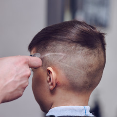 Obraz premium Barber is using clipper while making hair tattoo for a cute European boy in barbershop.