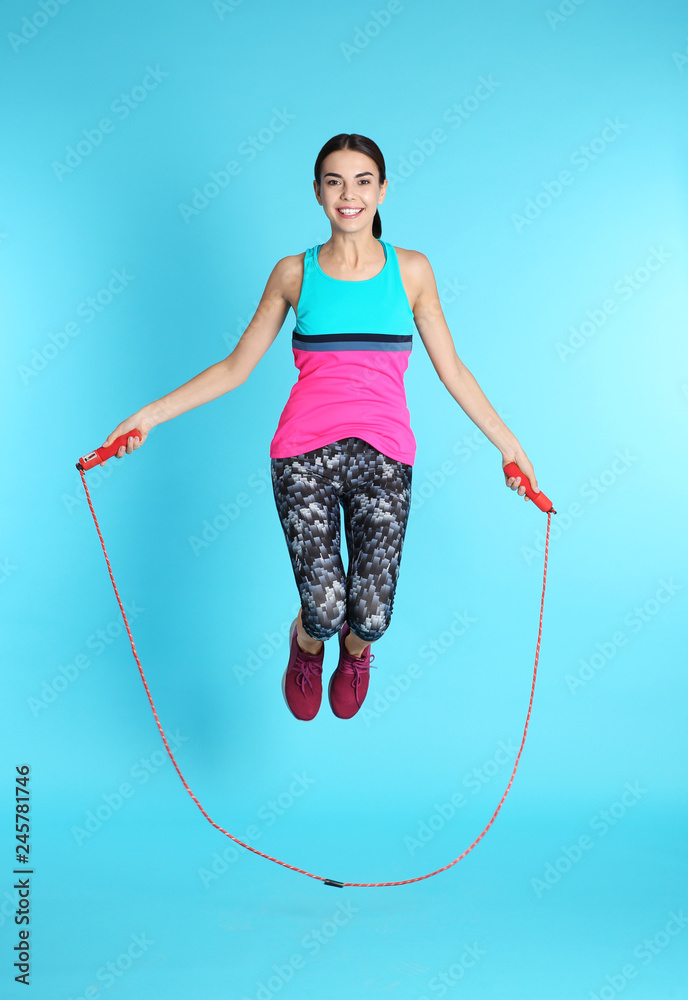 Wall mural Full length portrait of young sportive woman training with jump rope on color background