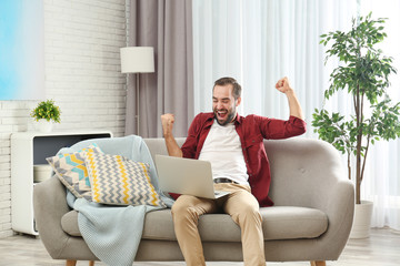 Obraz premium Emotional young man with laptop celebrating victory on sofa at home