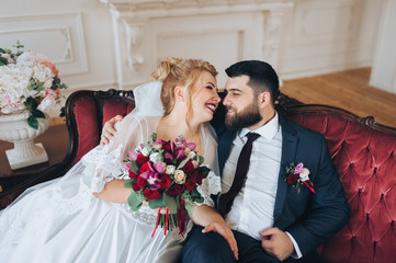 Stylish bearded groom and beautiful blonde bride hugging on a red armchair in a white studio. Portrait of lovers and smiling newlyweds in the interior. Wedding photography.