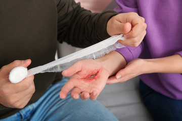 Young man applying bandage on woman's injured hand at home, closeup. First aid