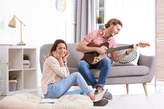 Young man playing electric guitar badly for displeased girlfriend in living room. Talentless musician