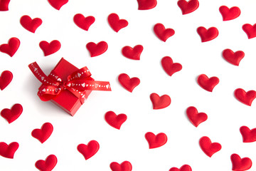 A red gift box with a heart-patterned ribbon sits among scattered red fabric hearts on a white background. Concept of: Heartfelt gift