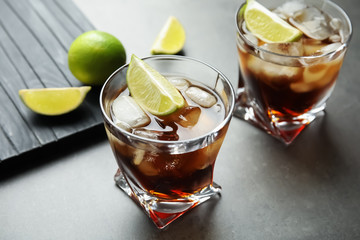 Glasses of cocktail with cola, ice and cut lime on grey background