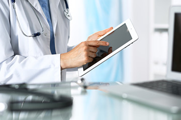 Fototapeta premium Woman doctor using tablet computer while sitting. Reflecting glass table is a working place of physician. Healthcare, insurance and medicine concept