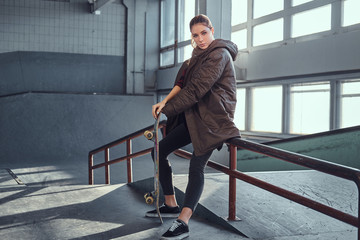 A beautiful young girl wearing a coat with her skateboard sitting on a grind rail in skatepark indoors.