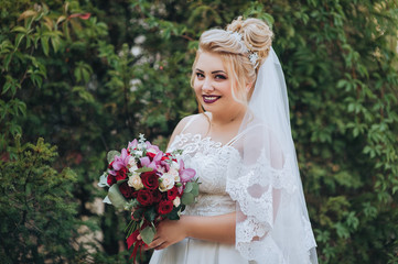 Closeup portrait of a beautiful blonde bride with a bouquet of roses in greenery and in a park. Wedding photography. Smiling and cute bride.