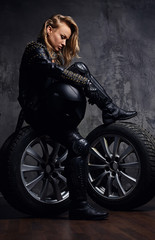Obraz na płótnie Canvas Fashion, sport, extreme. Beautiful woman wearing racer uniform, posing in a studio while sitting on a wheel and holding a protective helmet.