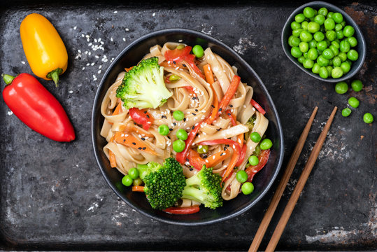 Vegetable stir fry with udon noodles, broccoli, carrot, pepper and green peas in bowl on black background. Top view
