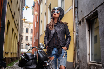 Girl in protective helmet and sunglasses wearing a leather jacket and ripped jeans standing near a black scooter on a old narrow street.