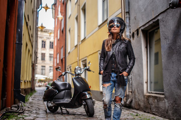 Fototapeta na wymiar Girl in protective helmet and sunglasses wearing a leather jacket and ripped jeans standing near a black scooter on a old narrow street.