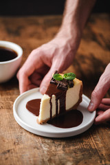 Cheesecake with chocolate sauce and cup of coffee. Hands holding slice of chocolate cake on plate
