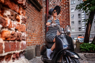 Fototapeta na wymiar Woman wearing dress and hat sitting on a black classic Italian scooter on an old street in Europe.