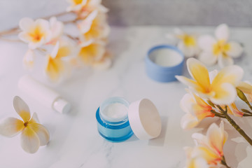 group of skincare products including moisturiser scrub and hand cream pots on marble table with exotic frangipani flowers