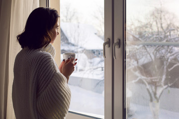 girl in a white knitted sweater stands at the window