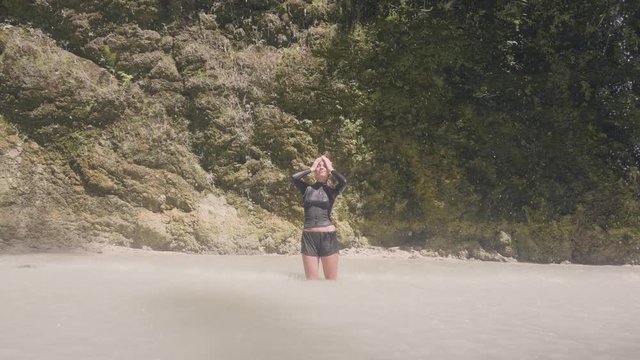 Young woman in wet clothes enjoying flowing waterfall in tropical forest. Happy woman standing under splashing water stream from mountain waterfall. Travel in wild jungle.