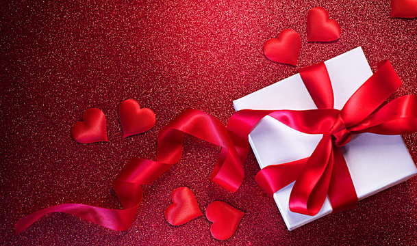 Valentine's Day romantic background with gift box and red satin hearts. Gift box over holiday red glittering background with hearts. Top view, flatlay