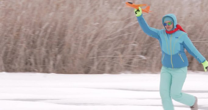 Young happy woman in red scarf and blue ski suit with orange plane with glider in her hands runs along icy snow-covered river. Bad weather, storm warning, village, winter, snow