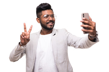 Smiling indian hipster man taking selfie with mobile phone with peace sign in eyeglasses with beard standing on white background.