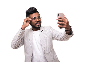 Smiling indian hipster man taking selfie with mobile phone in eyeglasses with beard standing on white background.