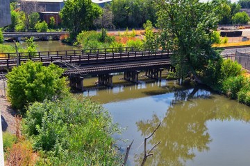 Fototapeta premium Views of Jordan River Trail Pedestrian and Train Track Bridge with surrounding trees, Russian Olive, cottonwood and muddy stream along the Wasatch Front Rocky Mountains, in Salt Lake City, Utah.