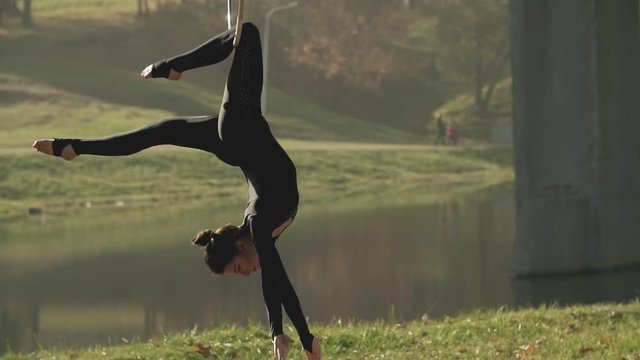 Air gymnastics woman performs acrobatics tricks on aerial hoop. Girl doing some acrobatic elements on aerial hoop outdoors. Flexible brunette with bare feet hanging in ring for aerial acrobatics