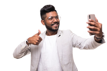 Smiling indian hipster man taking selfie with mobile phone with thumbs up in eyeglasses with beard standing on white background.
