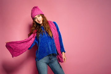 Modern young woman wearing blue sweater and pink hat, and scarf posing, making funny facial expression. Retouched, studio portrait.