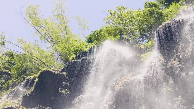 Splashing water from mountain waterfall in tropical forest slow motion. View from bottom water strea of flowing waterfall in rainforest in sunshine. Mountain river flowing on cliff in tropical forest.