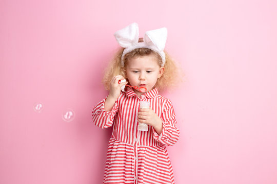 Lovely little girl in a striped red and white dress and bunny ears on her head inflates soap bubbles standing against a pink wall
