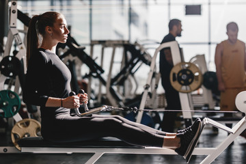 The beautiful athletic girl dressed dressed in a black sportswear is doing sport exercises with equipment on the bench in the modern gym