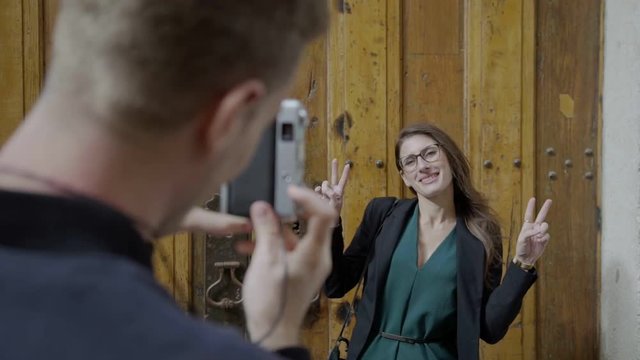 Smiling young woman in eyeglasses showing peace signs while standing near wooden door. back view of young man taking pictures with vintage camera of his girfriend. Photoshoot concept 