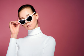 Fashion studio image of gorgeous elegant woman in white knitted golf and sunglasses posing over pink wall.