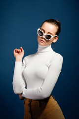 Portrait of a beautiful young girl in white knitted golf and sunglasses on a blue background